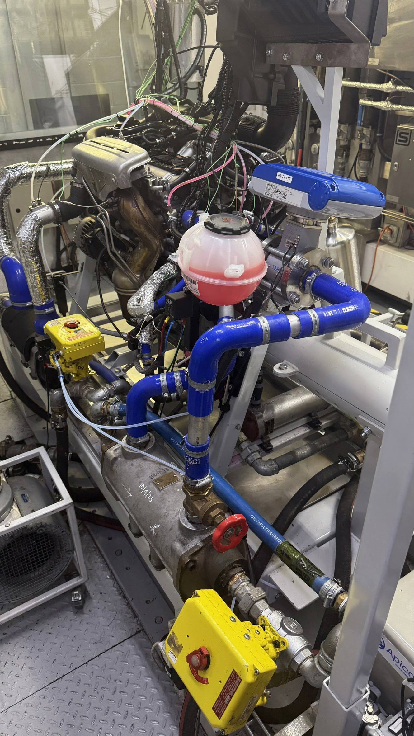 A close side view of an engine installed on a test bed, with multiple cables, sensors, insulated pipes, and coolant hoses connected for measurement. Industrial equipment, ventilation ducting, and control units surround the setup inside the test cell.