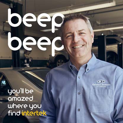 A technician with a Christian Broters Automotive uniform smiling inside a repair shop with vehicles in the background